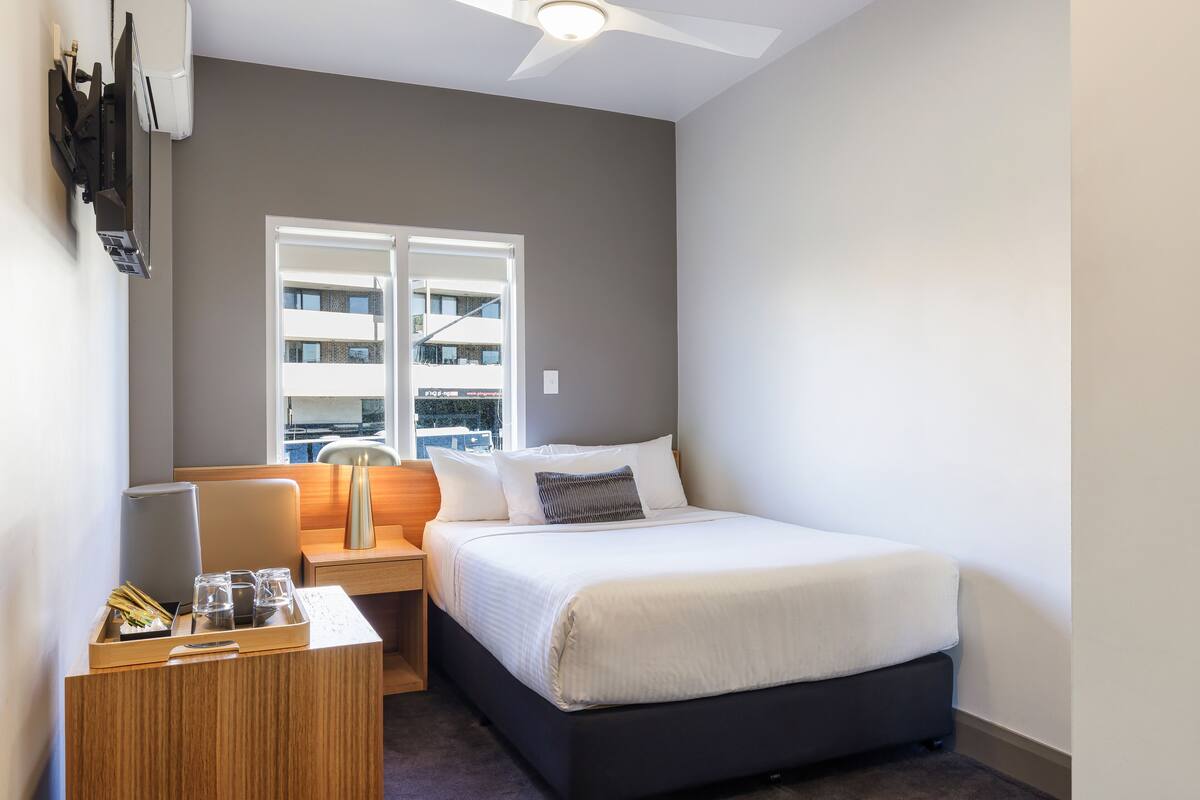 Une chambre d’hôtel équipée d’un lit, d’un bureau, d’une chaise et d’une fenêtre donnant sur des immeubles.