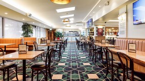 A hotel lobby with a geometric patterned carpet, wooden tables and chairs, orange booths, and a bar area in the background. - Kingsgrove Hotel (Kingsgrove)