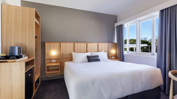 A hotel room with a large bed, wooden wardrobe, bedside table, and a view of the outdoors through the window. - Kingsgrove Hotel (Kingsgrove)