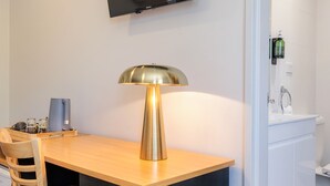 A hotel room with a wooden desk, a black TV mounted on the wall, a modern lamp, a chair, a small table with a coffee maker, and a bathroom visible through an open door. - Kingsgrove Hotel (Kingsgrove)