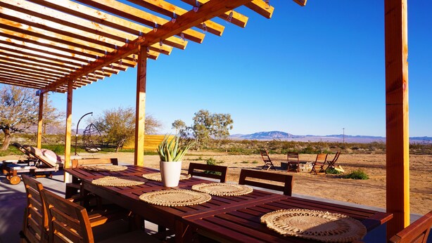 Une pergola en bois installée à l’extérieur, avec une table dressée pour les repas, entourée d’un paysage désertique et de montagnes.
