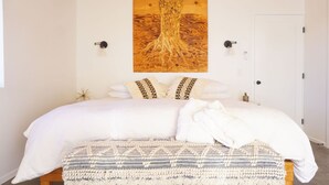 Un lit bien fait, avec du linge de lit blanc et un plaid à motifs, une œuvre d’art représentant un arbre accrochée au mur, et une porte blanche.