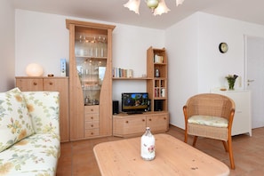 Ein Wohnzimmer mit einer Couch, einem Couchtisch, einem Fernseher, einem Bücherregal und einem Korbstuhl.