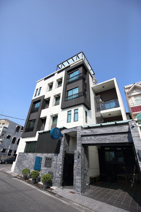 Um edifício moderno de vários andares com varandas e janelas, entrada de garagem e uma pequena área externa com mesas e cadeiras.