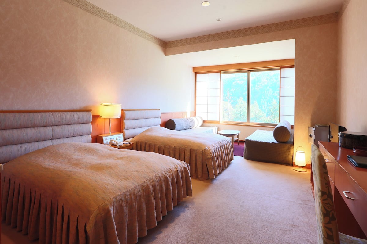 A hotel room with two beds, a desk, a chair, and a window with a view of trees.