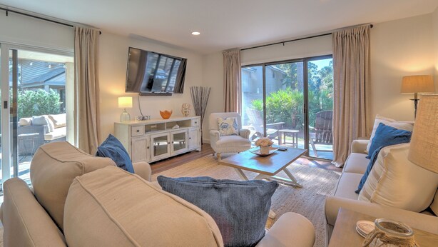 A spacious living room with a sofa set, a coffee table, a TV mounted on the wall, and a sliding glass door leading to an outdoor area. - SHIPYARD - WALK TO BEACH & STEPS TO POOL.  Bright corner unit & golf course view (Hilton Head Island)
