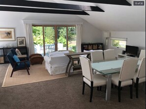 A modern living room with a glass dining table, white upholstered chairs, a blue armchair, and a sofa. There is a TV and a bookshelf in the room. - Resort Style Home- Family Friendly (Shelter Island)