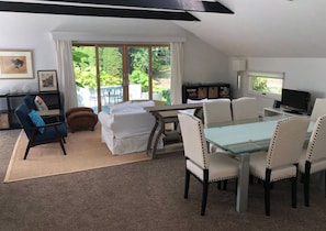 A modern living room with a glass dining table, white upholstered chairs, a blue armchair, and a sofa. There is a TV and a bookshelf in the room.
