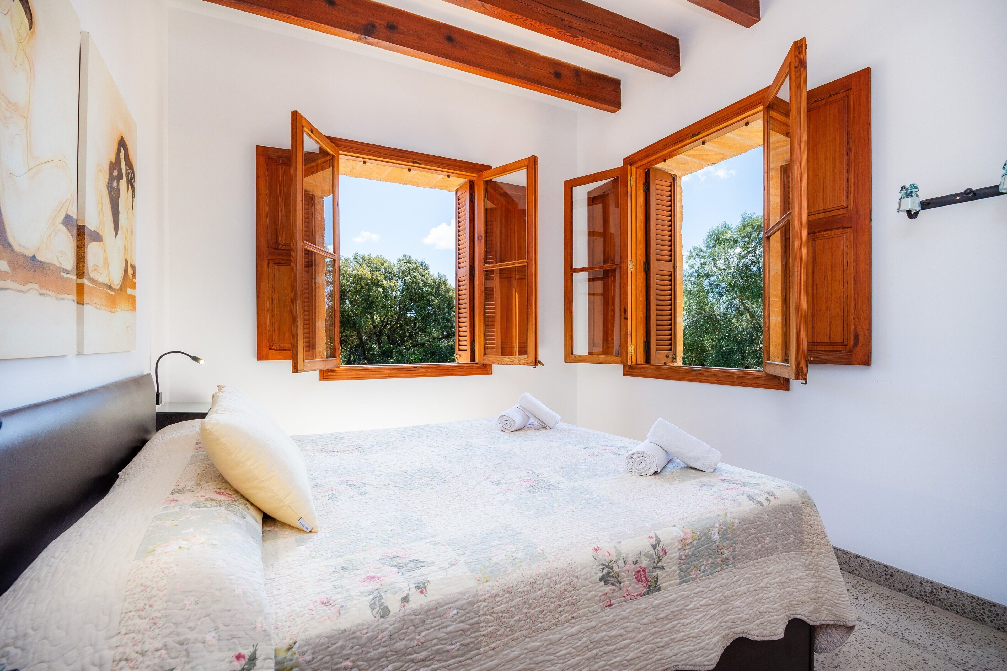 A bedroom with a bed, wooden shutters, a painting, and a window with a view of trees.