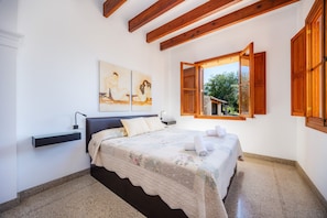 A bedroom with a bed, a nightstand, a wall-mounted shelf, a window with wooden shutters, and a painting on the wall. - Rustikale Finca mit Pool, Garten, WLAN, Klimaanlage und Balkon; Parkplätze vorhanden (Arta)