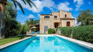 A stone house with a swimming pool, surrounded by greenery and palm trees.