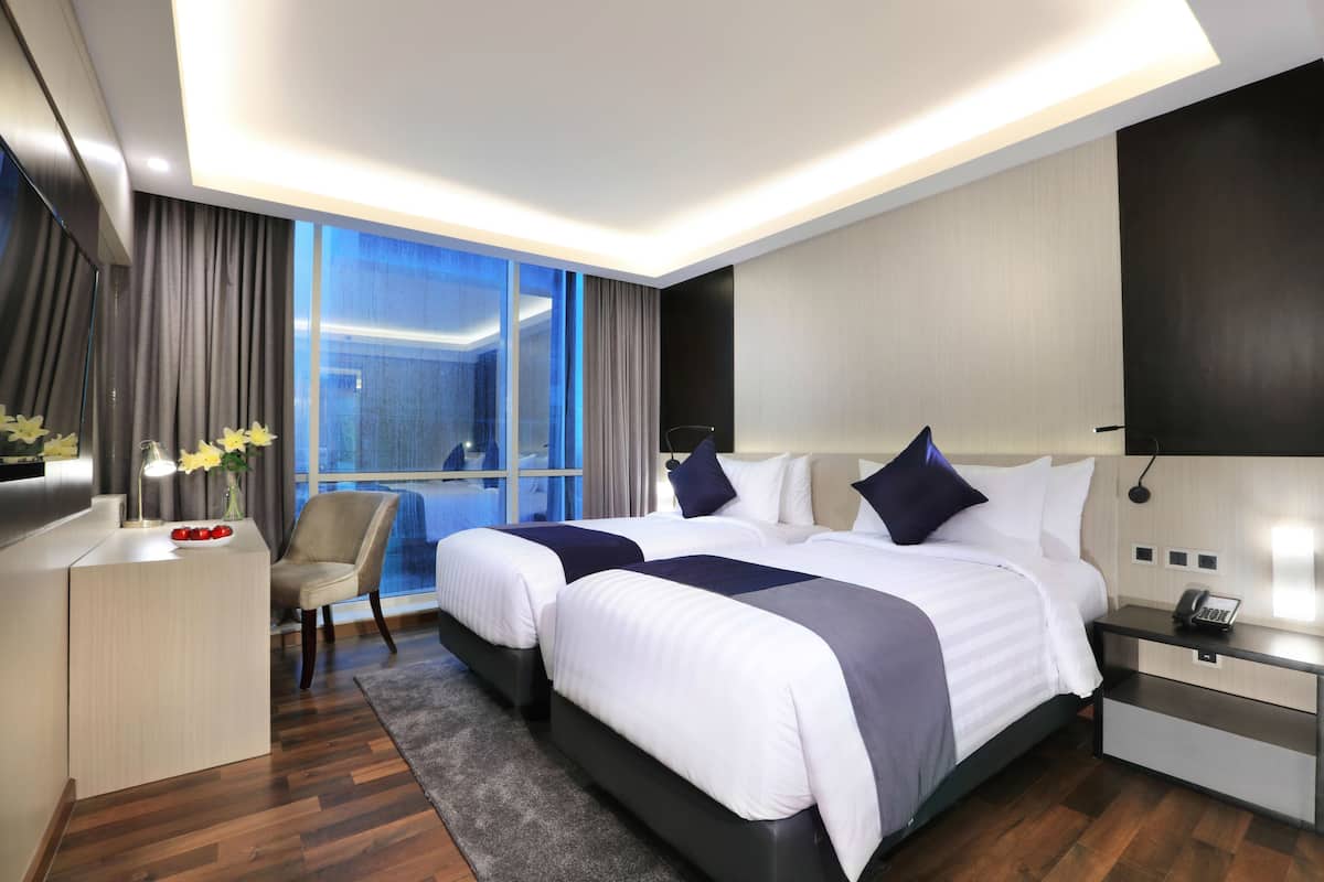 Een moderne hotelkamer met twee bedden, een bureau, een stoel, een nachtkastje en een groot raam met gordijnen.