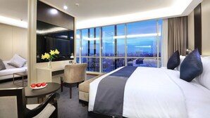 Een hotelkamer met een groot bed, een sofa, een bureau met een schaal fruit, een stoel en een groot raam met uitzicht op de stad.