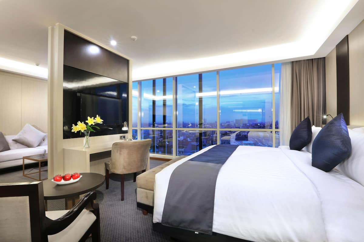Een hotelkamer met een groot bed, een sofa, een bureau met een schaal fruit, een stoel en een groot raam met uitzicht op de stad.