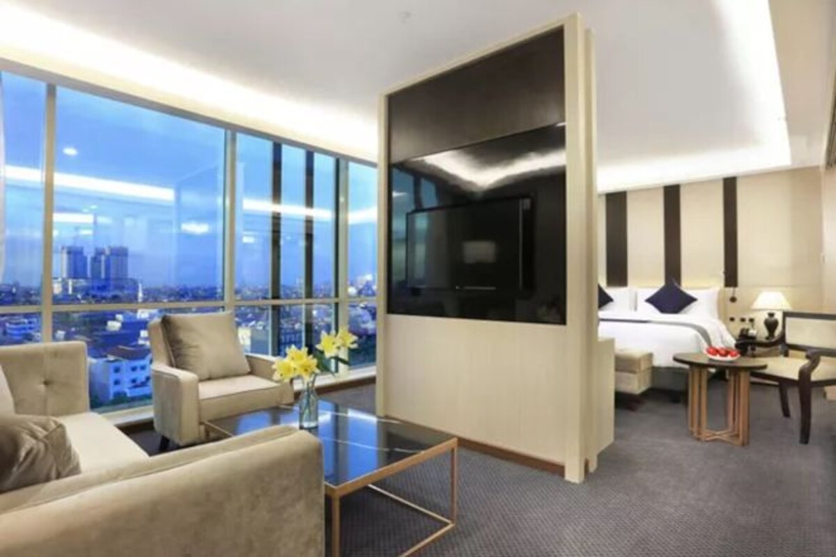 A hotel room with a large window, a bed, a desk, and a seating area with a view of the city.