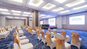A conference room with rows of white chairs with yellow sashes, a stage with a screen displaying 'ASTON,' and a blue carpeted floor.