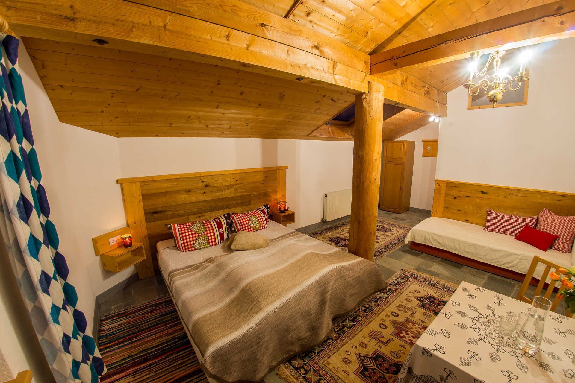 Une pièce chaleureuse avec un grand lit, un plus petit lit, des poutres en bois, une table avec des verres et un tapis à motifs.