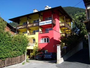 Ein mehrstöckiges Gebäude mit Balkonen, mit der Aufschrift 'Europa Pension', Fassade in Rot und Gelb, parkende Autos und einem Bergpanorama im Hintergrund.