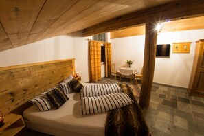 Een knus vertrek met een houten plafond, een bed met gestreepte kussens, een klein tafeltje met een vaas en een televisie aan de muur.