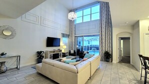 A spacious living room with a large window, a sofa, a TV, and a dining area. - Voluminous * 5 Bd Pool/Spa Home in Champions Gate (Davenport)
