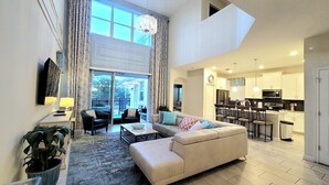 A modern living room with a sofa, coffee table, and dining area. A kitchen with white cabinets and a dining table is visible in the background. - Voluminous * 5 Bd Pool/Spa Home in Champions Gate (Davenport)