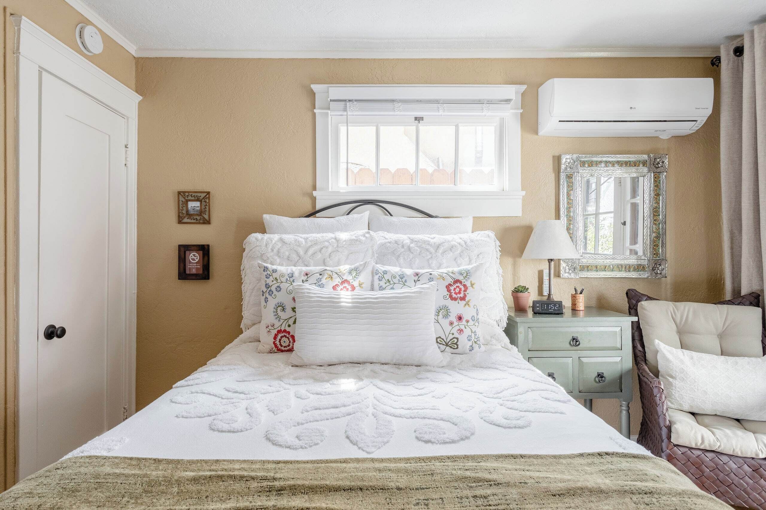 A bedroom with a bed, a window, an air conditioner, a bedside table, and a mirror.