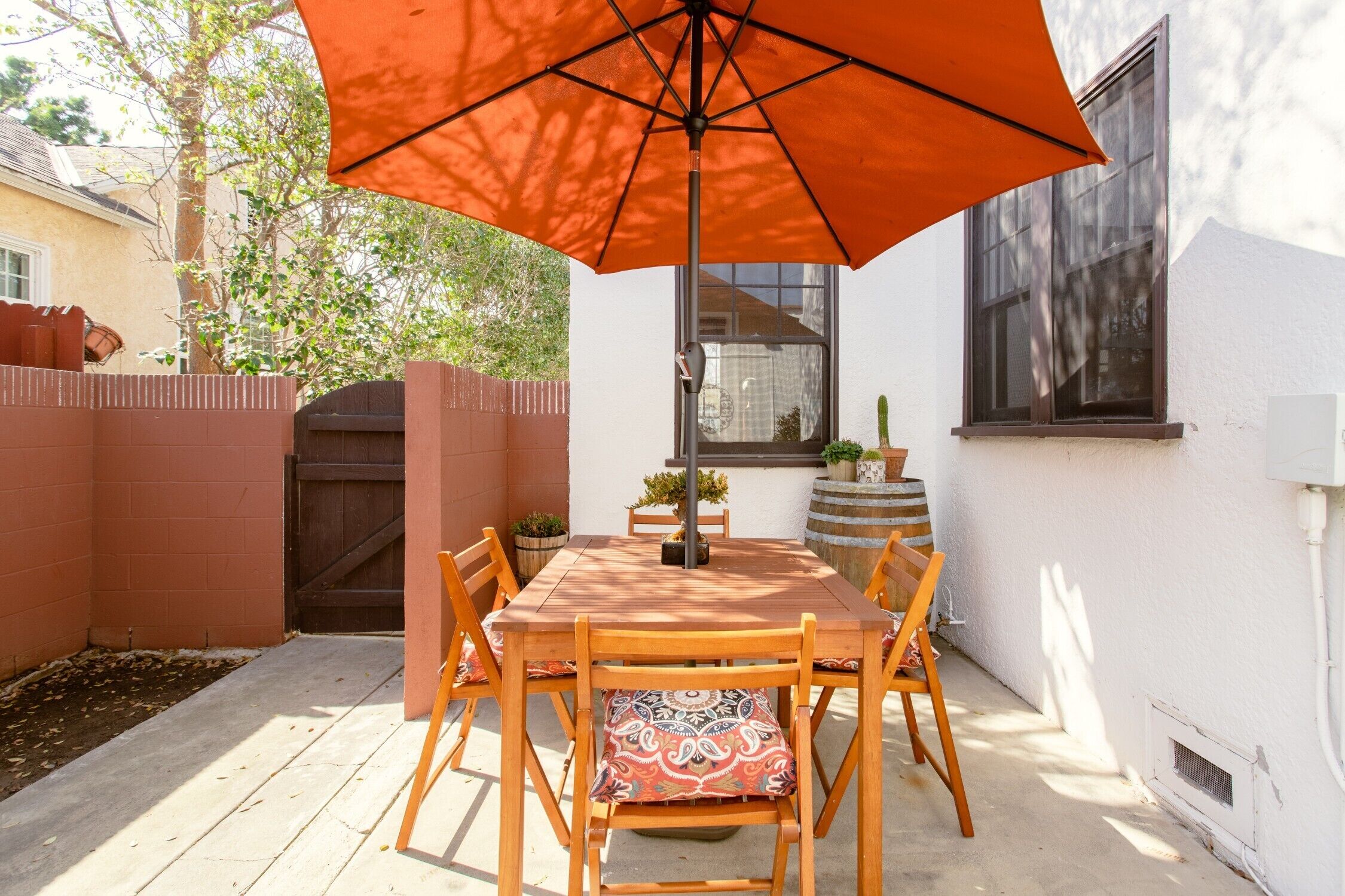 A patio with a wooden table and chairs, an orange umbrella, and a potted plant.