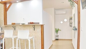 A small kitchenette with two white bar stools, a wooden ceiling, and a white wall with a shelf holding a coffee pot and cups.