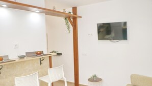 A modern kitchen with a wooden bar, white chairs, a TV mounted on the wall, and a small side table with a plant.
