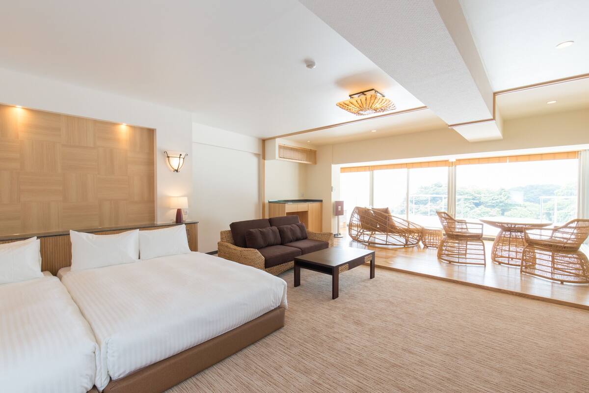 A hotel room with a large bed, a sofa, a coffee table, wicker chairs, and a view of the outdoors.