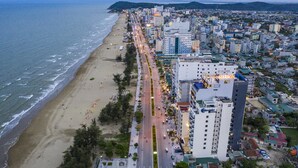 A coastal cityscape with a beach, a main road, and several buildings including a prominent hotel with a sign. - The Marron Hotel (Sam Son)