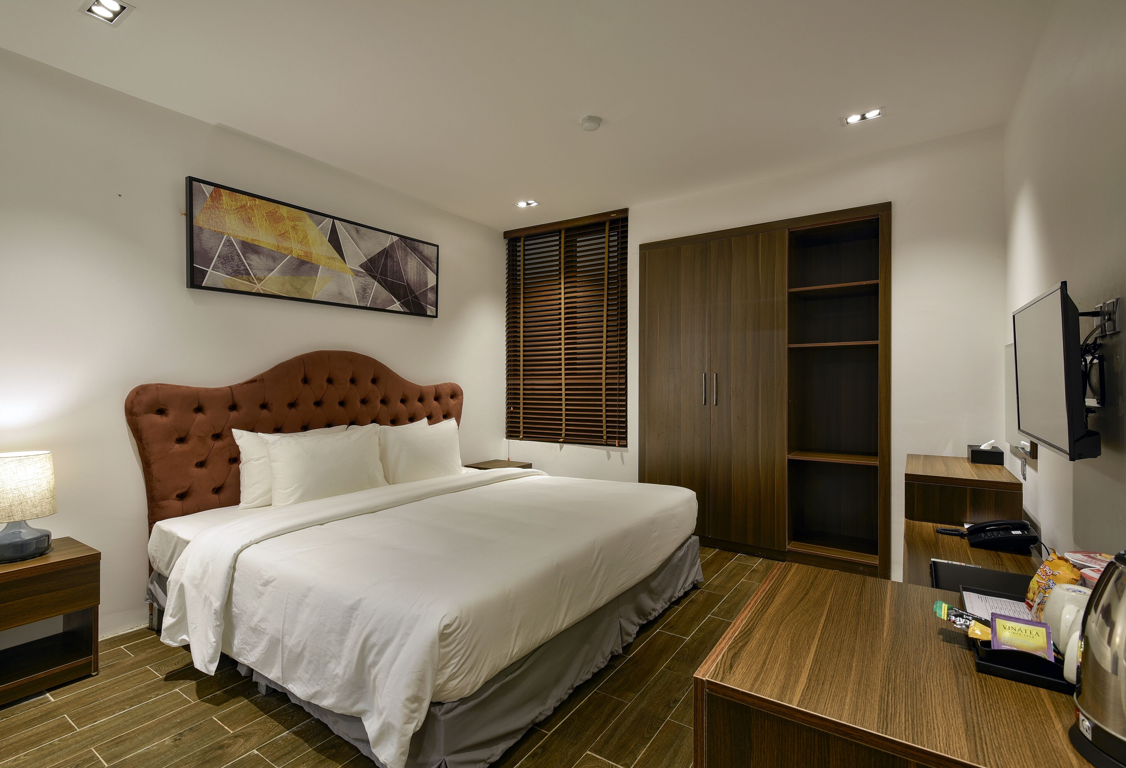 A modern hotel room with a large bed, a wooden wardrobe, a desk with a chair, and a mounted television.