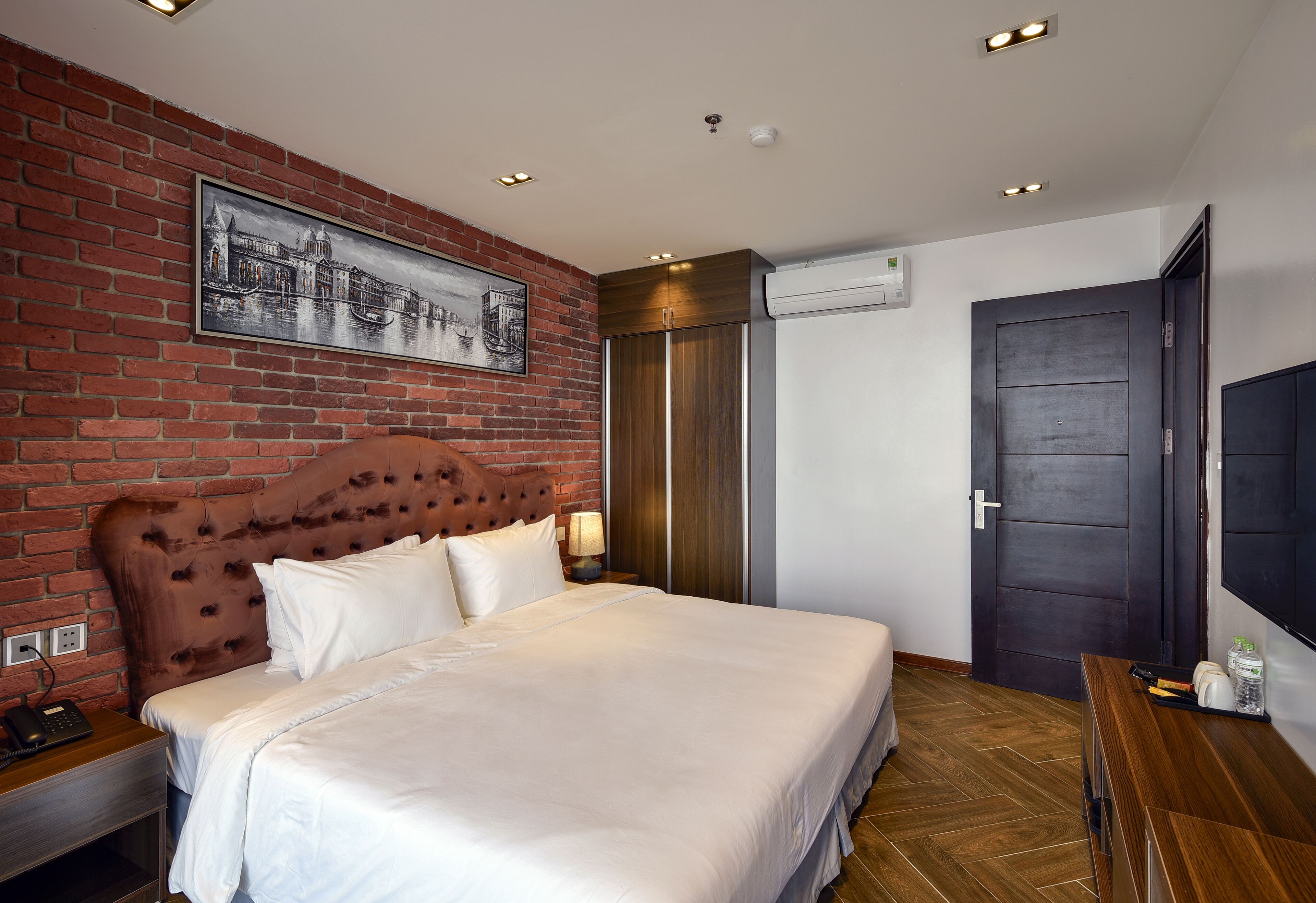 A hotel room with a brick wall, a large bed, a wooden headboard, a flat-screen TV, and a wardrobe.