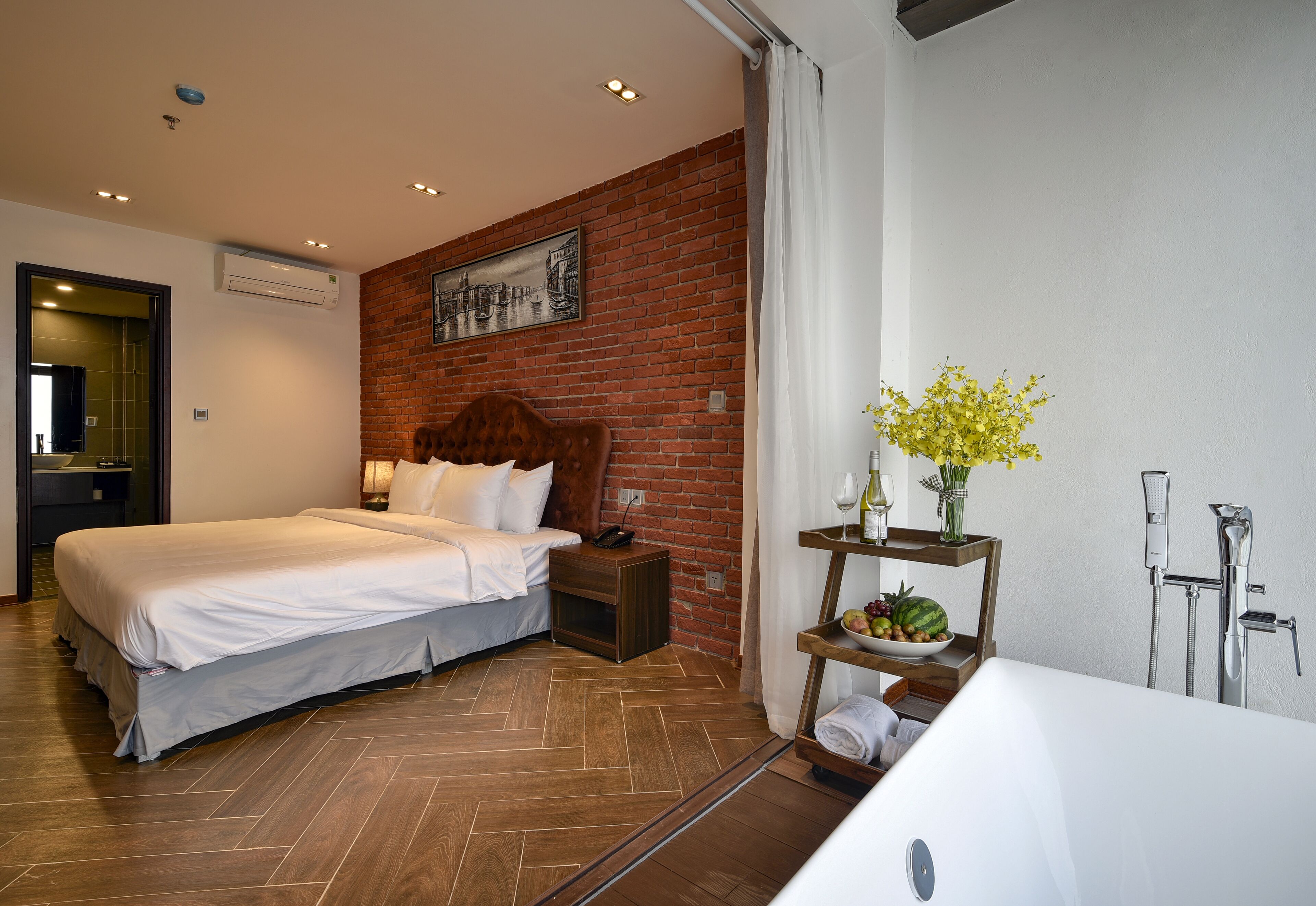 A hotel room with a bed, a wooden nightstand, a brick wall, a bathroom with a bathtub, and a small table with fruit and flowers.