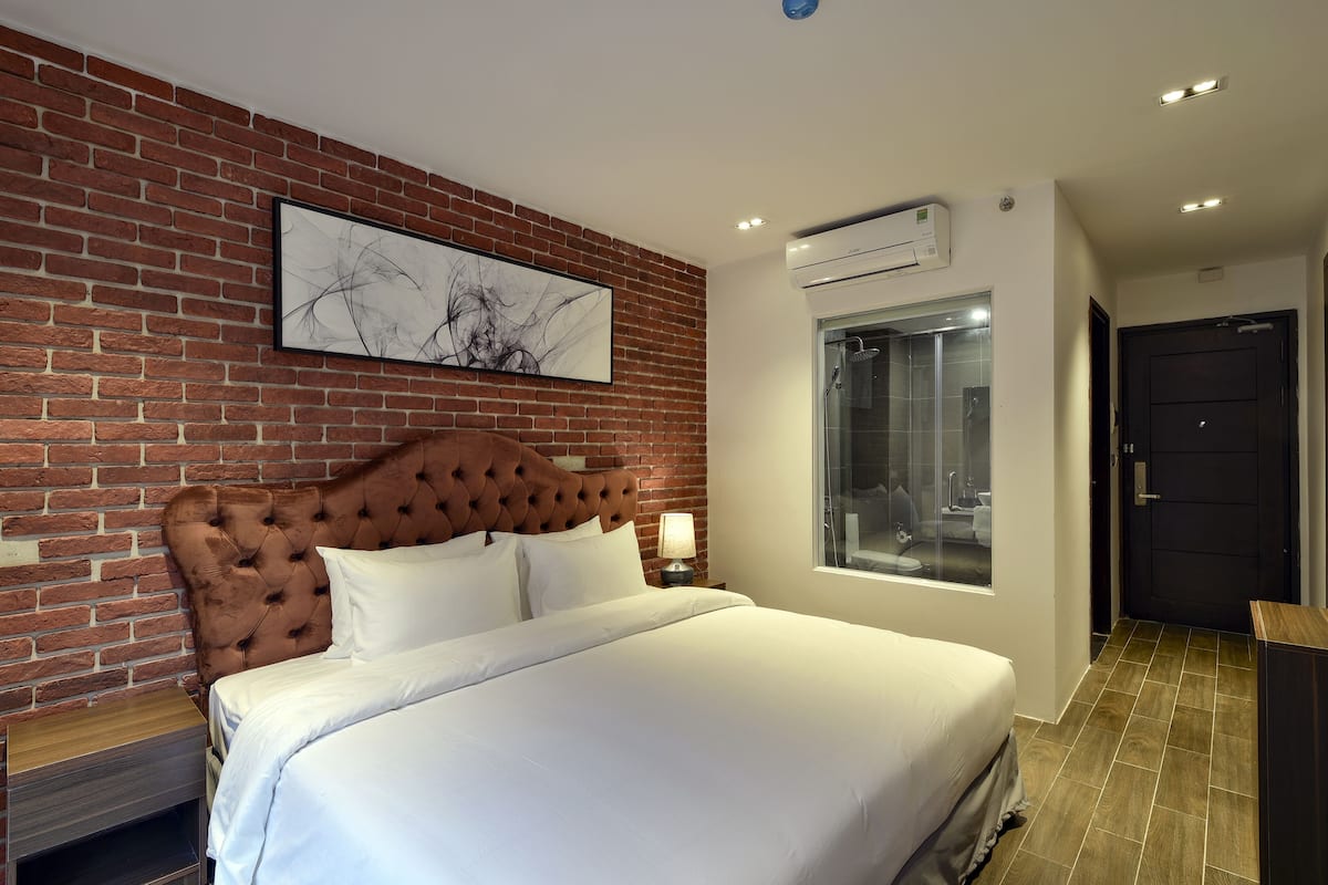 A modern hotel room with a brick wall, a tufted headboard, a large bed, a window with a view, and an air conditioning unit.