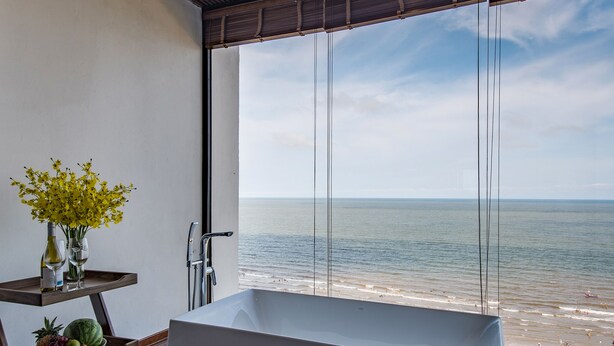 A modern bathroom with a large bathtub, a wooden shelf with towels and fruit, and a view of the beach. - The Marron Hotel (Sam Son)