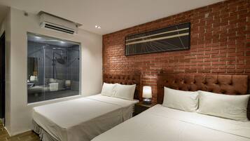 Habitación de hotel con dos camas, ducha con cerramiento de vidrio, pared de ladrillo y lámpara moderna.