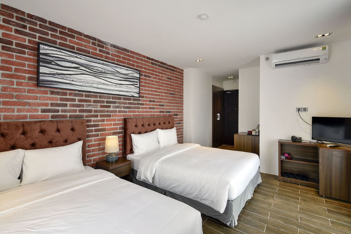 A hotel room with two beds, a brick wall, a mounted artwork, an air conditioning unit, a television, and a wooden cabinet.