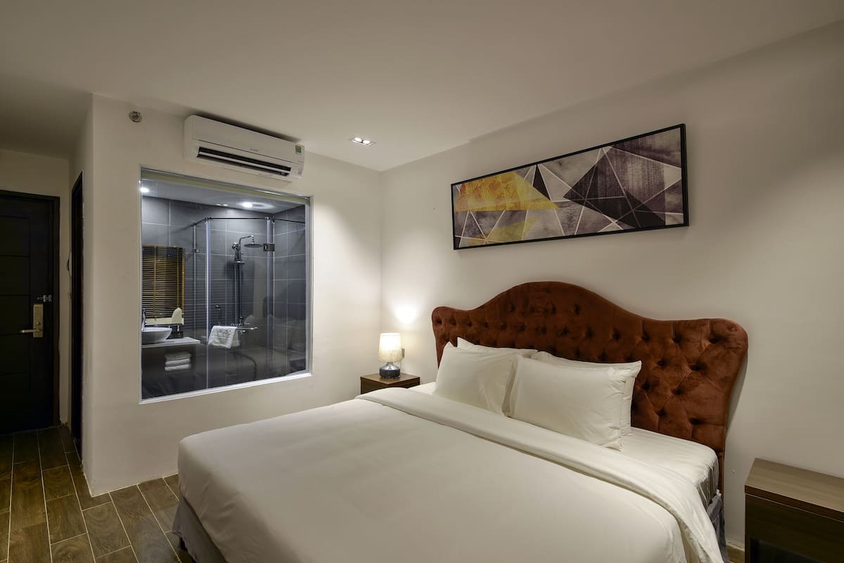 A hotel room with a large bed, a window with a view of a bathroom, a wall-mounted air conditioner, and a framed abstract painting.