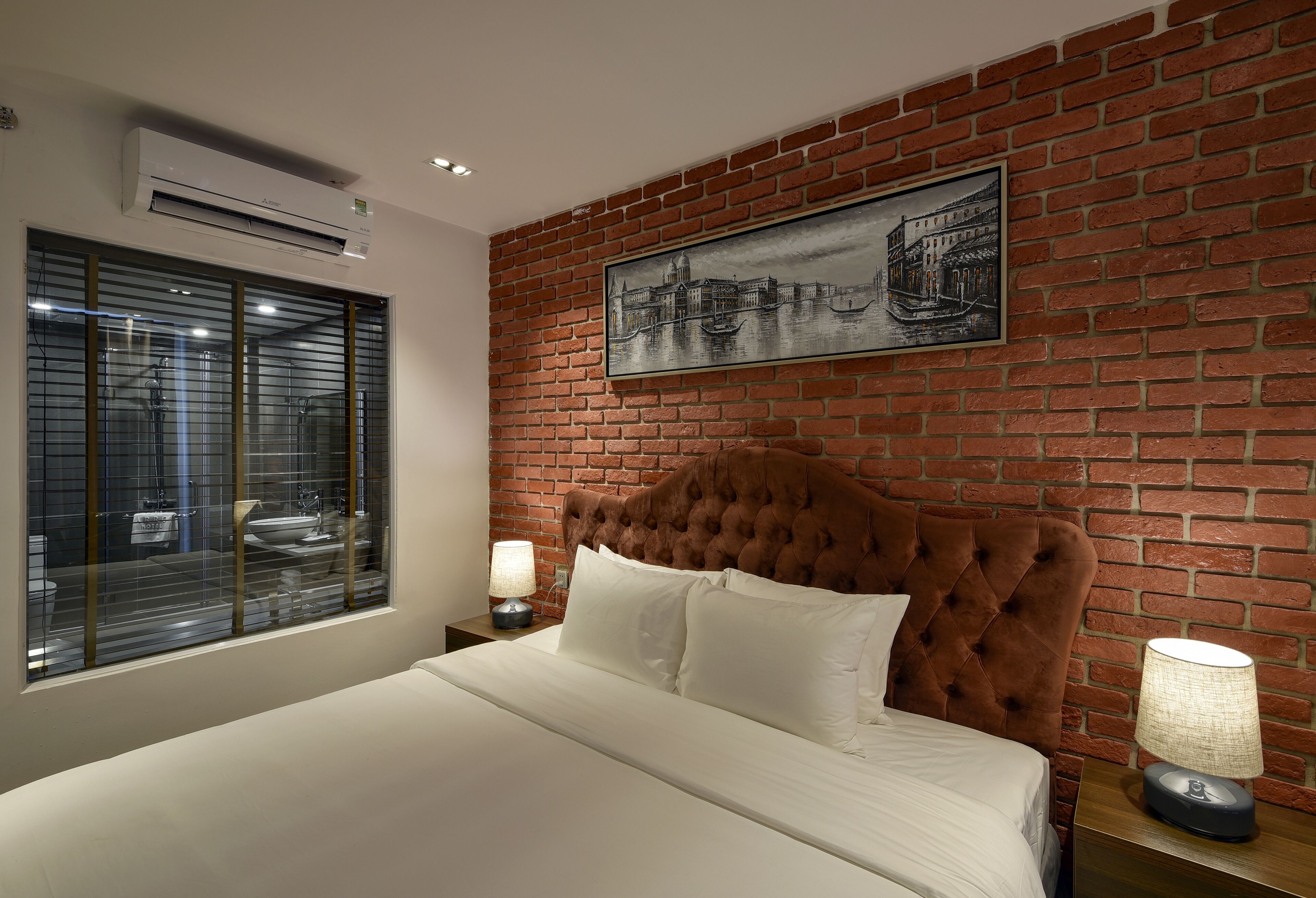 A bedroom with a brick wall, a large bed with white linens, and a window with blinds.