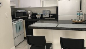 A modern kitchen with white cabinets, a microwave, a coffee maker, and a sink. There are two black bar stools and a small island with a paper towel holder.