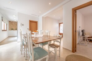 A dining area with a wooden table and white chairs, a kitchen in the background, and a white wall with a door.