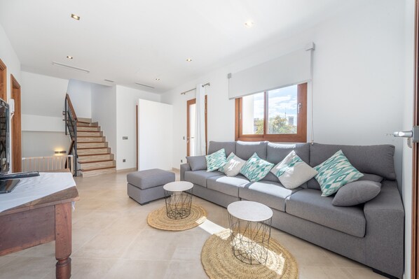 A spacious living area with a grey sofa, two round side tables, a wooden coffee table, and a staircase leading to an upper floor.