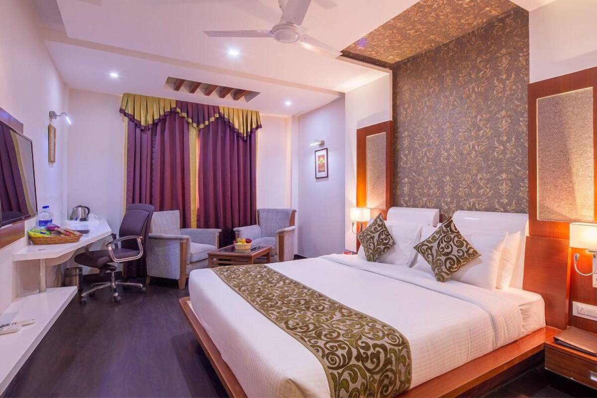 A hotel room with a bed, two chairs, a desk, a ceiling fan, and a bathroom mirror.