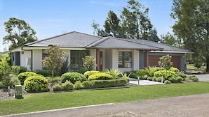A single-story house with a gabled roof, a front porch, and a well-maintained front yard with various shrubs and a small tree. - 1OAK - THE VINTAGE HOLIDAY HOUSES (POKOLBIN)