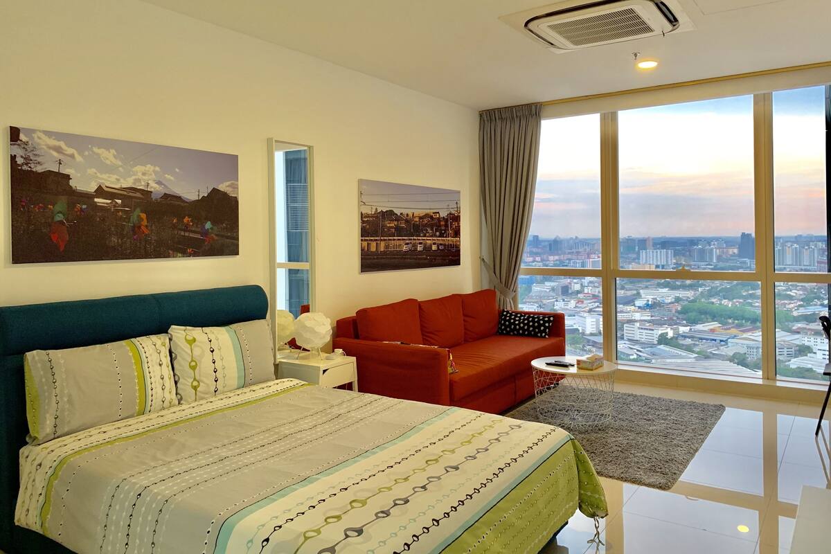 Quarto de hotel moderno com uma cama grande, um sofá vermelho e vista para a cidade através de amplas janelas.
