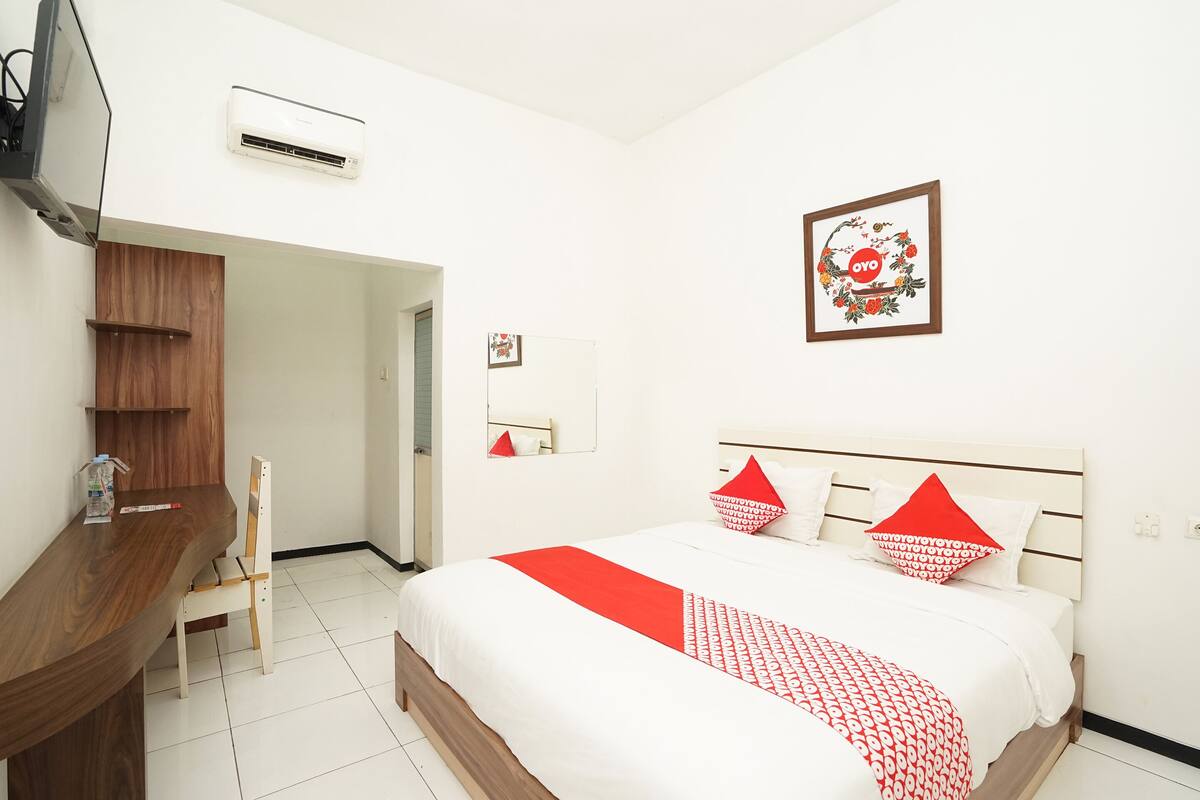 A hotel room with a bed, a wooden desk, a chair, a TV, an air conditioner, and a framed picture on the wall.