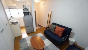 A compact living space with a kitchenette, a sofa, a coffee table, and a floor lamp. - Apartment Camon EBR0001E (Osaka)
