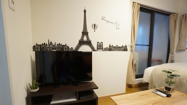 A hotel room with a large flat-screen TV on a wooden stand, a small potted plant, a remote control, and a bed with a beige cover. - Apartment Camon EBR0001E (Osaka)