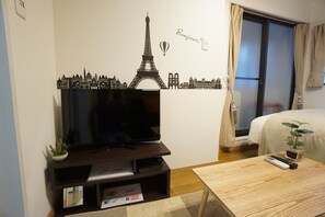 A hotel room with a large flat-screen TV on a wooden stand, a small potted plant, a remote control, and a bed with a beige cover. - Apartment Camon EBR0001E (Osaka)
